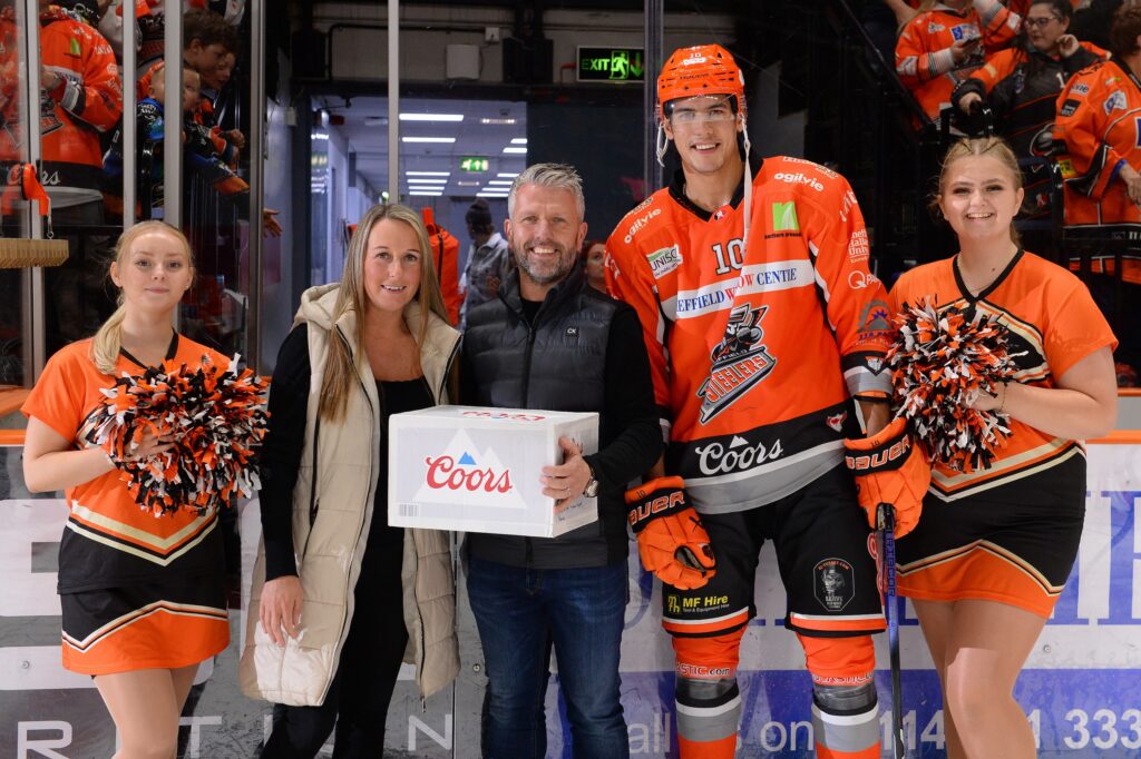 The image shows an ice hockey player, cheerleaders and a fan receiving a prize won in a raffle